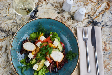 Greek salad with giant olives and provencal herbs served in blue ceramic plate with glass of white wine on marble table. Top view or flat lay.