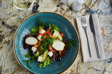 Greek salad with giant olives and provencal herbs served in blue ceramic plate with glass of white wine on marble table. Top view or flat lay.