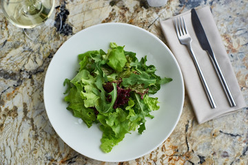 Lettuce salad served in white bowl with glass of white wine. Knife and fork lying on textile napkin on marble table. Top view or flat lay.