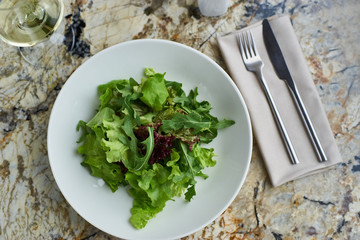 Lettuce salad served in white bowl with glass of white wine. Knife and fork lying on textile napkin on marble table. Top view or flat lay.