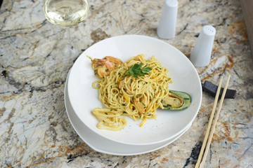 Flat egg noodles with seafood served in big white ceramic round plate with glass of white wine on marble table