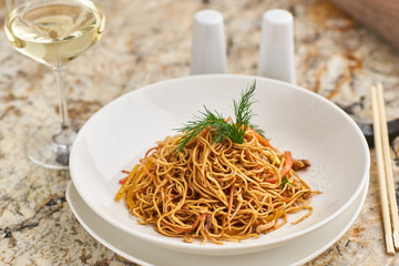 Thin egg noodles with vegetables and soy sauce served on white round plate with glass of white wine and chopsticks