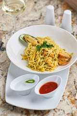 Flat egg noodles with seafood served in big white ceramic round plate with glass of white wine on marble table