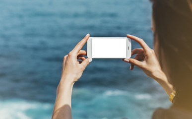 Hipster photograph on smart phone gadget mobile, mock up of blank screen. Traveler hold and using in female hand mobile on background mountain seascape horizon. Tourist look on blue ocean, lifestyle