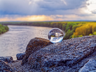 Magic glass ball on old stones. In the background is the bend of the river, spring forest and cloudy sky - obrazy, fototapety, plakaty