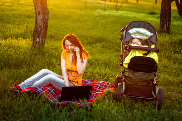 beautiful a red hair woman using computer and talking on smart phone sitting on grass in outdoor park on the sunset with sleeping baby in carrige