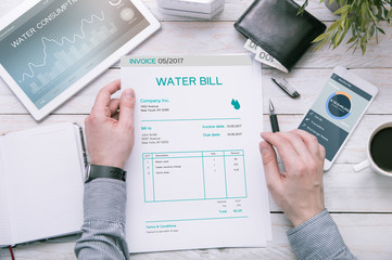 Man holds invoice of water usage over desk with tablet and smartphone with applications made in...
