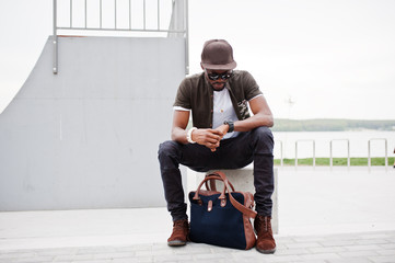 Portrait of sitting stylish african american man wear on sunglasses and cap, looking on his watches...