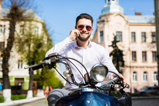 Cheerful young man sitting on scooter and talking on the mobile phone