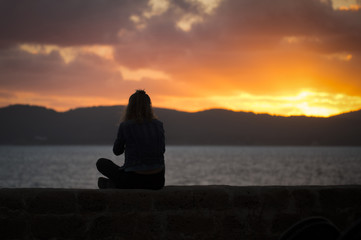Fototapeta premium A silhouette of a girl is relaxing at sunset enjoying the view of the sea from the ancient walls of a fortification in Alghero at sunset,