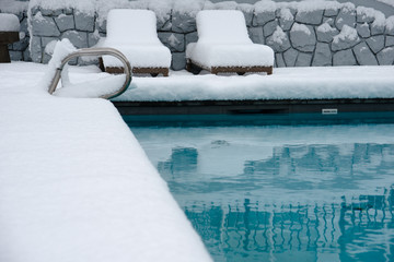An outdoor swimming pool. the area around it and the lounging chairs  covered in over half a foot, 20cm, of snow.