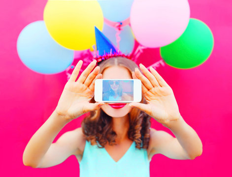 Display of phone woman in a birthday cap is taking a picture on a smartphone with an air colorful balloons on a pink background