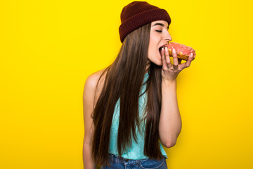 Hungry funny girl in hat biting pink donut near yellow wall