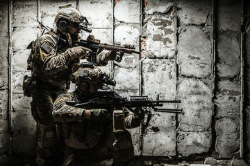 Pair of Army Rangers with rifle and machine gun moving along the concrete wall on mission. They are ready to start firing if enemy appear. Outdoor location shot