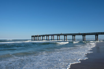 Obraz premium St Johns County Ocean Pier stretching to the horizon, sea lapping on the sandy shore of the beach in St Augustine, Florida, USA on a sunny afternoon. Clear blue sky with no clouds, St Augustine Beach.