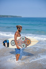 Full-length vertical portrait of an attractive bearded curly man with a surfboard is standing on a...