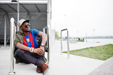 Fototapeta na wymiar Stylish african american boy wear at cap, football t-shirt and sunglasses sitting near steel railings. Black sports man portrait.