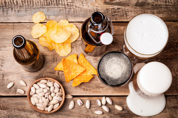 Beer in bottles and glasses on a wooden table. Beer and snacks are pistachio nuts, chips and nachos top view. Drink and snack for the football match or party.