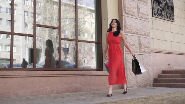 young beautiful girl is walking down the city street with shopping bags after a successful shopping. Brunette in a dress, sunglasses and high-heeled shoes. Slow motion