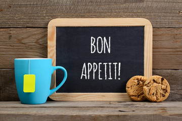 Wishes bon appetit on small blackboard, cup of tea and cookies on wooden background