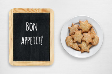 Cookies in plate and small blackboard with Bon appetit wishes on white wooden table
