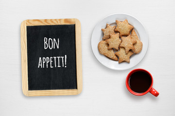 Cookies, coffee cup and small blackboard with Bon appetit wishes on white table