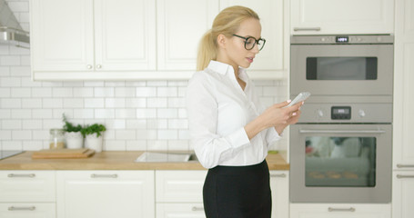 Beautiful successful businesswoman wearing official clothing and glasses using smart phone while standing at modern light kitchen.