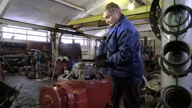 Professional senior mechanic repairs the rear axle of a dump truck. Heavy machine workshop. Worker moves the tractor gearbox on the telpher in the background.