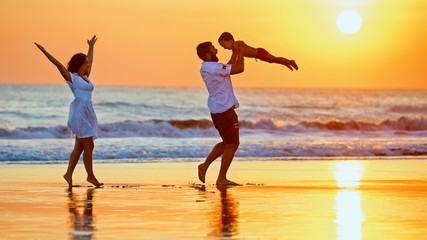 Happy family - father, mother, baby son walk with fun along edge of sunset sea surf on black sand...