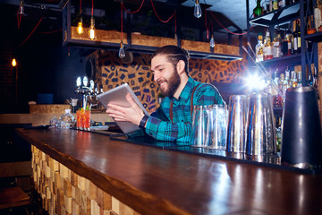 Barman with a hipster beard looks at  laptop behind  counter in the bar.