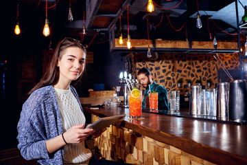 A girl with  cocktail smiles with tablet behind counter at the bar.