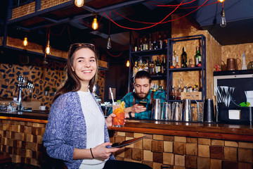 A girl with  cocktail smiles with tablet behind counter at the bar.