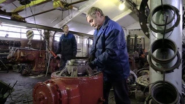 Professional senior mechanic repairs the rear axle of a dump truck. Heavy machine workshop. Worker moves the tractor gearbox on the telpher in the background.