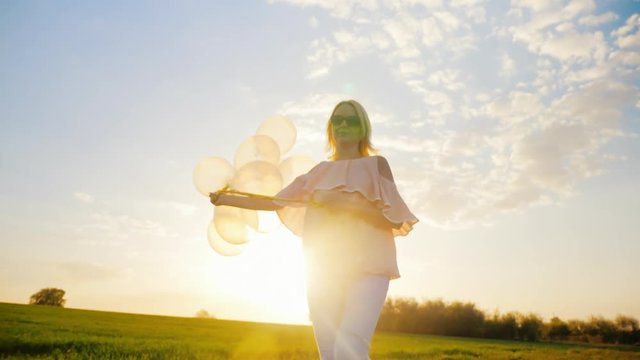 Carefree young woman with balloons walking on a green meadow at sunset. Concept - lightness and freedom of movement, dream. Slow motion video