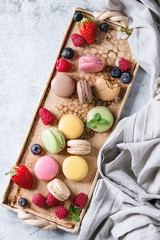 Variety of colorful french sweet dessert macaron macaroons with different fillings served on terracotta tray with spring flowers, berries, textile over gray texture background. Top view with space
