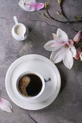 White cup of black coffee, served on white saucer with jug of cream and magnolia flower blossom branch over gray texture background. Flat lay, space