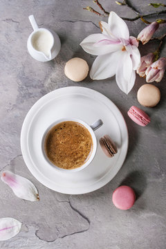 White cup of black coffee, served on white saucer with jug of cream, macaroons biscuits and magnolia flower blossom branch over gray texture background. Flat lay, space