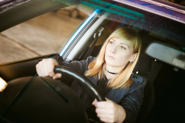 Portrait blonde woman sitting behind wheel in driver's seat of blue car. Hands on handlebars, driving problems of female