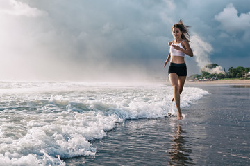 Active sporty woman run along ocean surf by water pool to keep fit and health. Sunset black sand beach background with sun. Woman fitness, jogging training and sport activity on summer family holiday.
