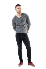 Young handsome casual man with hands behind back smiling at camera. Full body length portrait isolated over white studio background.