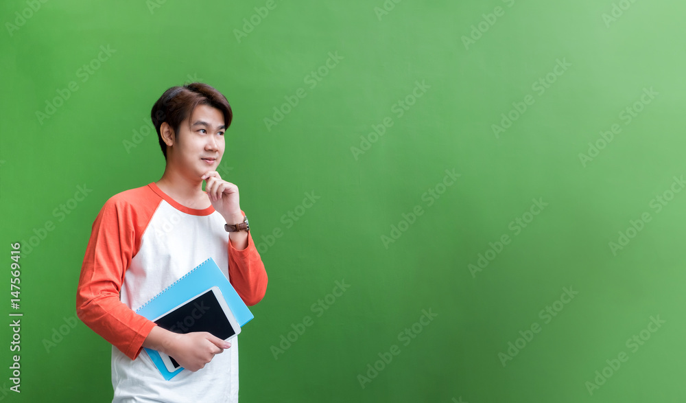 Poster young teenager man holding tablet computer and notebook and thinking action at green wall in univers