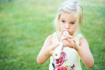 Blond child girl playing flute outdoors in the garden with green grass lawn background © Petr Bonek