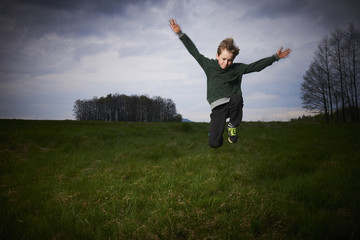 Child blond boy jumping in a spring green meadow against a storm's cloudy sky at sunset
