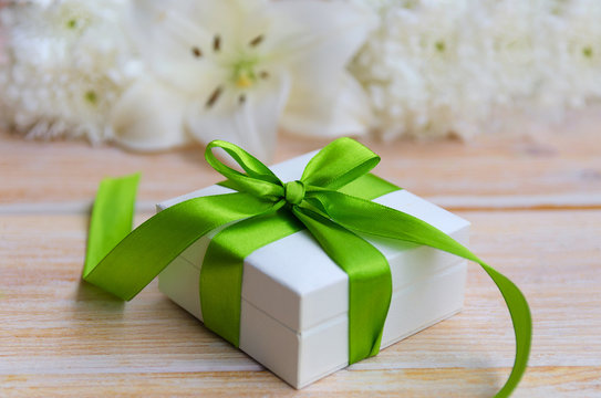 White lilie flower and white gift box with green ribbon and bow on light wooden background. Beautiful greeting card . Happy Mothers day or Happy birthday concept.