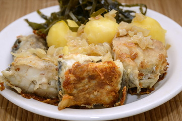 Fried fish, boiled potatoes and sea kale lies on a plate on a bamboo napkin. Close-up, selective focus.