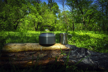 Fototapeta premium Kettle and mug on the charred log in sunny spring day with forest and tent background