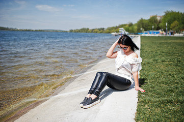 Brunette girl on women's leather pants and white blouse, sunglasses, sitting on quay of lake.
