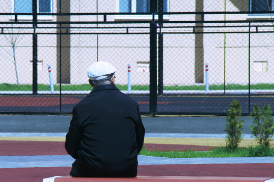 A lonely old man sits on a bench with his back to us and looks at the sports basketball court. A symbol of old age, loneliness, ill health and the desire to become young again