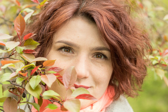 Pretty romantic young red head woman with apple branches red leaves in spring park. Spring trees background. Portrait close up.