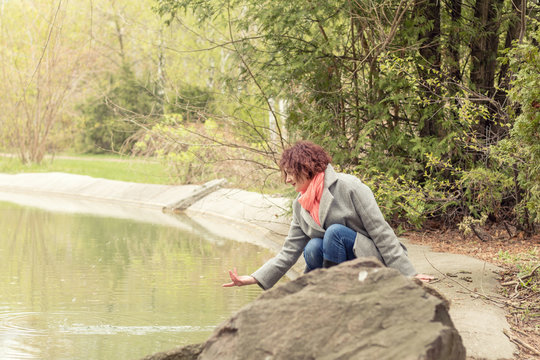 Pretty romantic young red head woman sitting close to the pond in spring park. Spring trees background.
